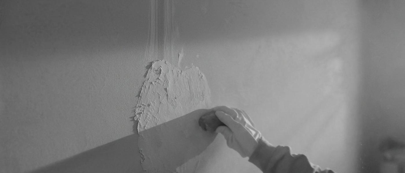 Gloved hands sanding a feathered drywall patch smooth with a fine sanding sponge in raking daylight