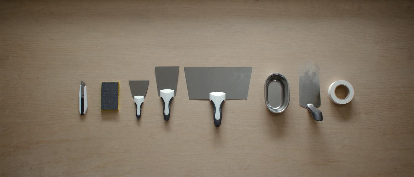 Drywall finishing tools laid out on plywood: taping knives, mud pan, corner trowel, paper tape and a sanding sponge