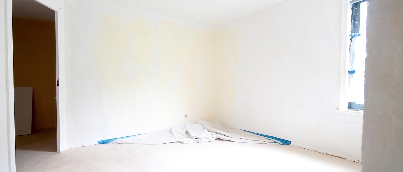 An empty apartment room mid-turnover with smooth repaired drywall in fresh primer, a folded drop cloth and painter's tape along the baseboard